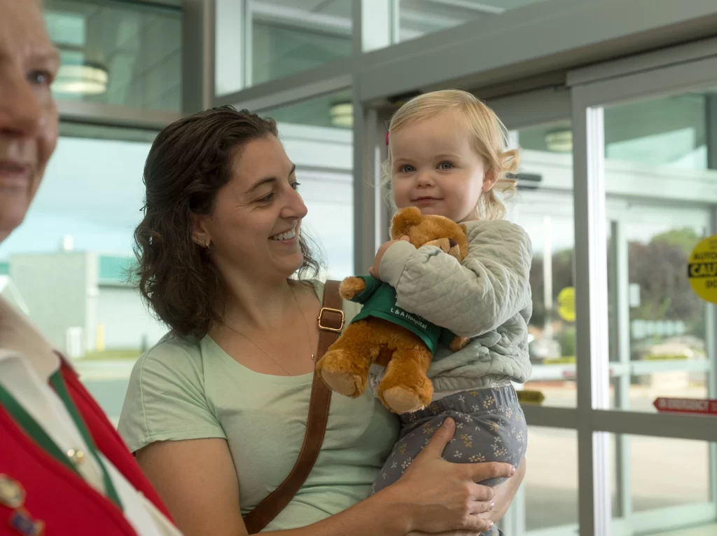 An adult holding a toddler indoors, with the child holding a stuffed toy and looking toward the camera.