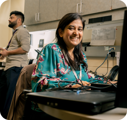 Staff member smiling while working at a computer workstation in an office, with a colleague working in the background.