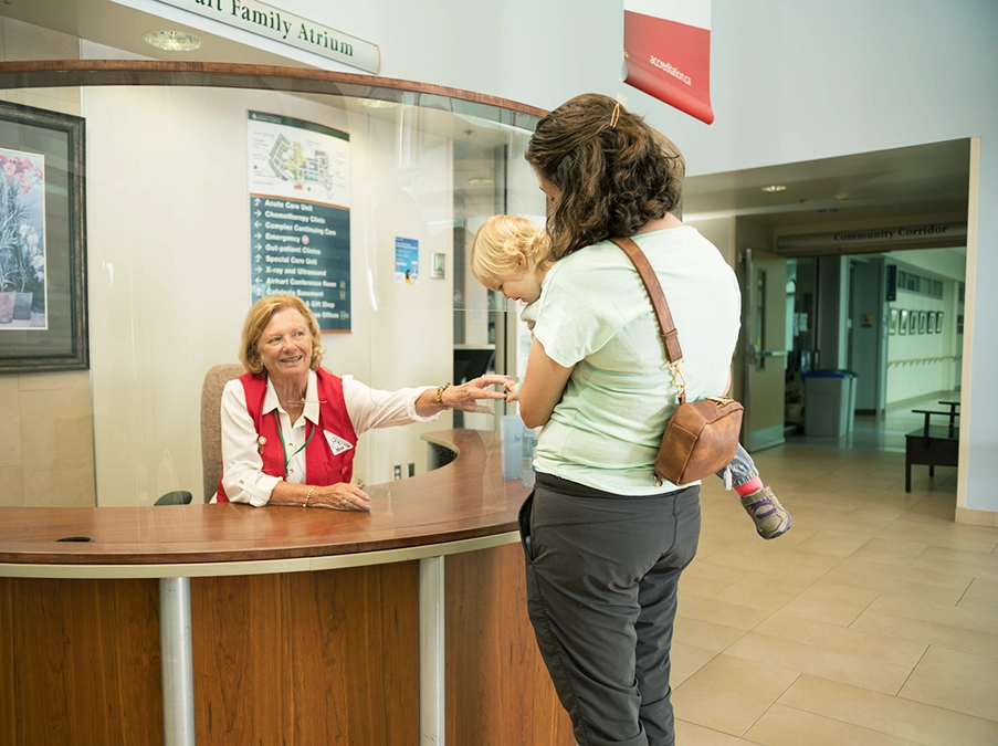 Hospital volunteer behind a reception desk speaks with a parent holding a young child in a hospital atrium.