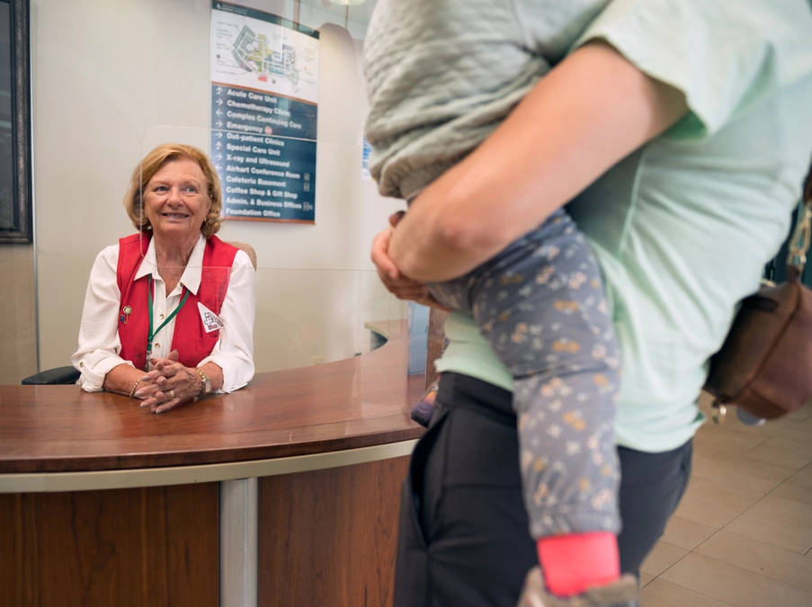 Hospital volunteer greets a parent holding a child at the front desk.