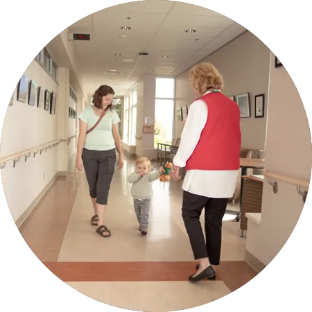 Hospital volunteer walks with a parent and young child in a hospital hallway, holding a teddy bear.
