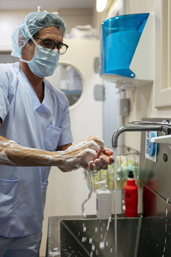 Healthcare worker wearing a mask and hair cover washes hands at a clinical sink.