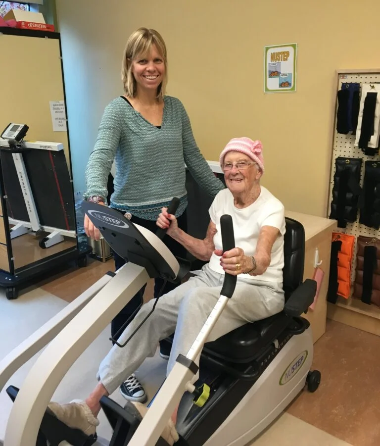 Resident using exercise equipment with staff support in a therapy room.